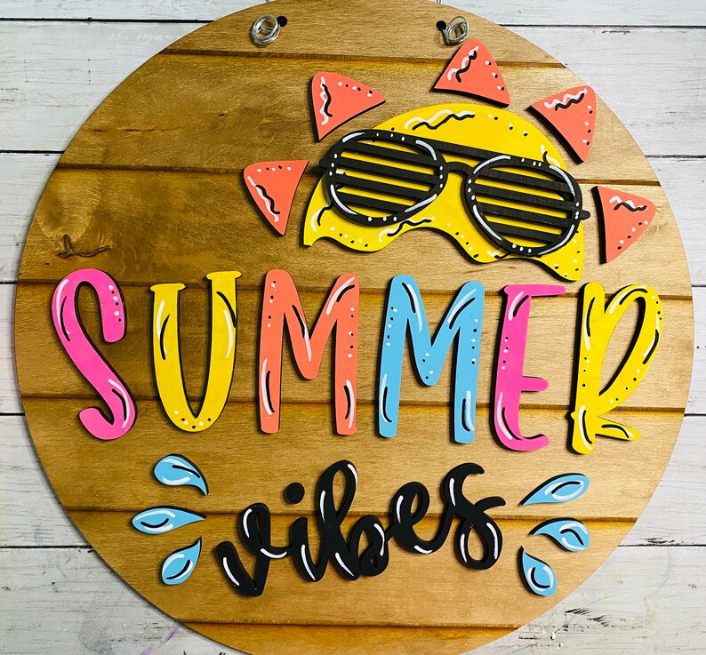 May include: A round wooden sign with the words "Summer Vibes" in colorful letters. A yellow sun with sunglasses is at the top of the sign.
