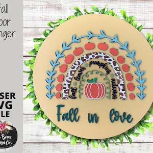 May include: A round wooden door hanger with a fall-themed design. It features a rainbow made of leaves, pumpkins, and the words "fall leaves", "pumpkins", and "autumn". The words "Fall in love" are written below the rainbow.