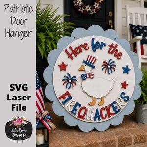 May include: A round wooden door hanger with a scalloped light blue edge, featuring a white centre with a duck wearing a patriotic top hat and bow tie. The text reads Here for the FIREQUACKERS, accented by red and blue stars and fireworks.