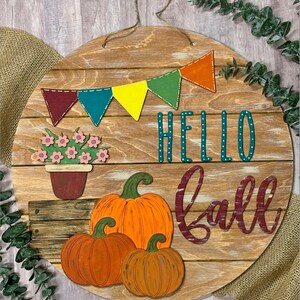 Puede incluir: Un letrero redondo de madera con las palabras "Hello Fall" en letras turquesa y burdeos. El letrero est&aacute; decorado con una hilera de banderines coloridos, una maceta con flores y tres calabazas naranjas.