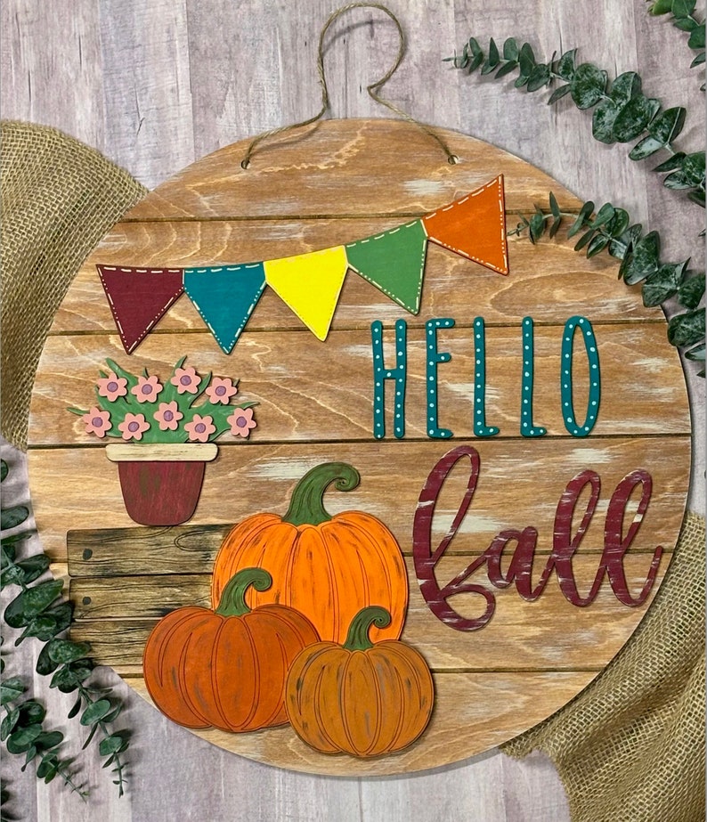 Puede incluir: Un letrero redondo de madera con las palabras "Hello Fall" en letras turquesa y burdeos. El letrero est&aacute; decorado con una hilera de banderines coloridos, una maceta con flores y tres calabazas naranjas.