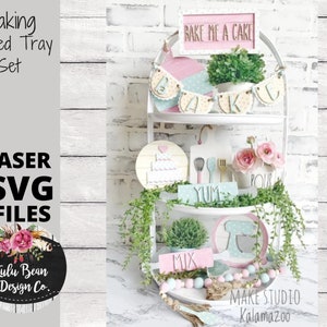 May include: A three-tiered white tray set with various baking-themed decorations. The top tier has a pink and white banner that says "Bake Me A Cake". The middle tier has a round wooden sign with a heart and a cake, and a wooden sign that says "Yum". The bottom tier has a round wooden sign with a mixer, and a wooden sign that says "Mix".