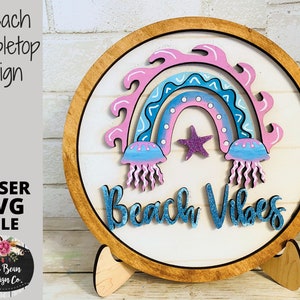 May include: A wooden tabletop sign with a pink, blue, and white rainbow, two jellyfish, and a star. The sign says "Beach Vibes" in blue glitter letters.