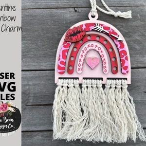 Op de afbeelding: Een roze en rode houten regenboog autohanger met een hart, lippen en een pijl. De regenboog heeft een franje van wit garen.