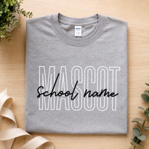 May include: A heather grey crew neck t-shirt featuring the word MASCOT in large white outlined capital letters with the words school name written in black cursive script across the centre. The shirt has a Gildan brand label at the neckline.