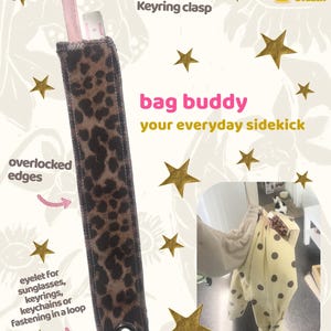 May include: A leopard print fabric strap featuring a metal keyring clasp at the top and a black metal eyelet at the bottom. The strap has visible overlocked edges and includes text reading bag buddy your everyday sidekick and Block & Bobbin.