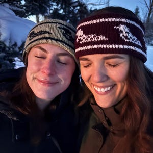 May include: Two people wearing winter knit hats and smiling. One hat is tan with a black design, the other is burgundy with a white design. The background shows snow and trees.
