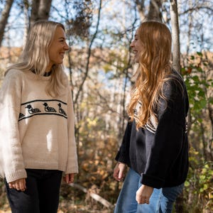 May include: Two people wearing sweaters stand in a forest setting. One sweater is cream-colored with a black duck design, and the other is black with a white and black pattern. Both are smiling. The background features trees and sunlight.