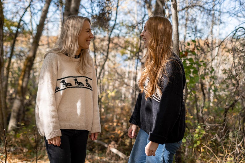 May include: Two people wearing sweaters stand in a forest setting. One sweater is cream-colored with a black duck design, and the other is black with a white and black pattern. Both are smiling. The background features trees and sunlight.