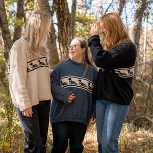 May include: Three people wearing sweaters with a horizontal band of loon bird designs. The sweaters are in cream, blue, and black. The people are standing in a wooded area with trees and foliage.