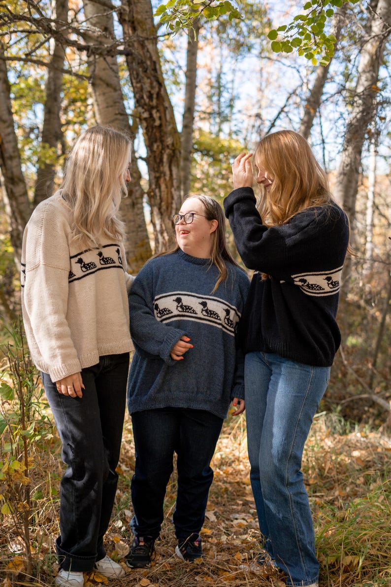 May include: Three people wearing sweaters with a horizontal band of loon bird designs. The sweaters are in cream, blue, and black. The people are standing in a wooded area with trees and foliage.