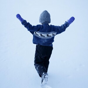 May include: A child in a blue sweater with a white pattern and gray knit hat, wearing blue mittens, walks in the snow with arms outstretched. The child is wearing black pants and black boots. The background is a snowy landscape.