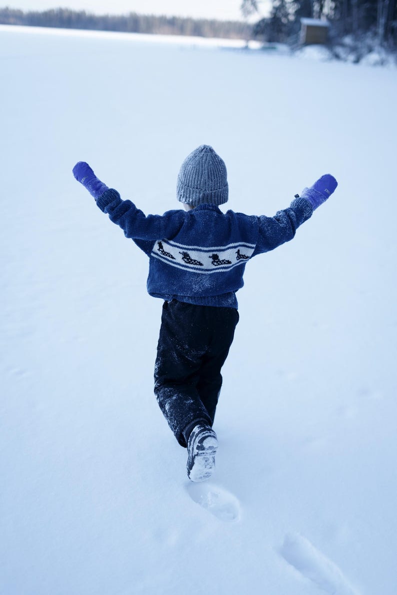 May include: A child in a blue sweater with a white pattern and gray knit hat, wearing blue mittens, walks in the snow with arms outstretched. The child is wearing black pants and black boots. The background is a snowy landscape.