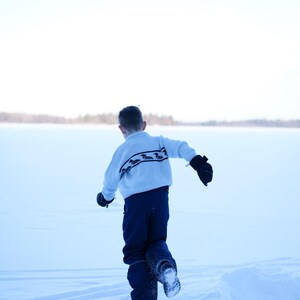 May include: A child wearing a light blue sweater with a black and white pattern, navy blue pants, and black mittens runs through a snowy landscape. The background features a snow-covered expanse and a distant treeline.