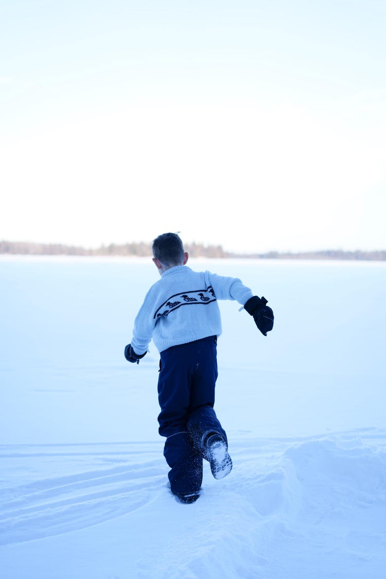 May include: A child wearing a light blue sweater with a black and white pattern, navy blue pants, and black mittens runs through a snowy landscape. The background features a snow-covered expanse and a distant treeline.