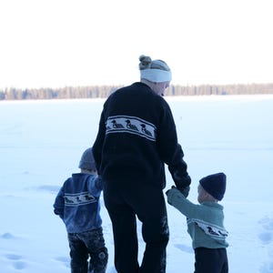 May include: A person and two children walking in the snow. The person wears a black sweater with a white and black pattern. The children wear sweaters and hats. The scene is set in a snowy landscape.