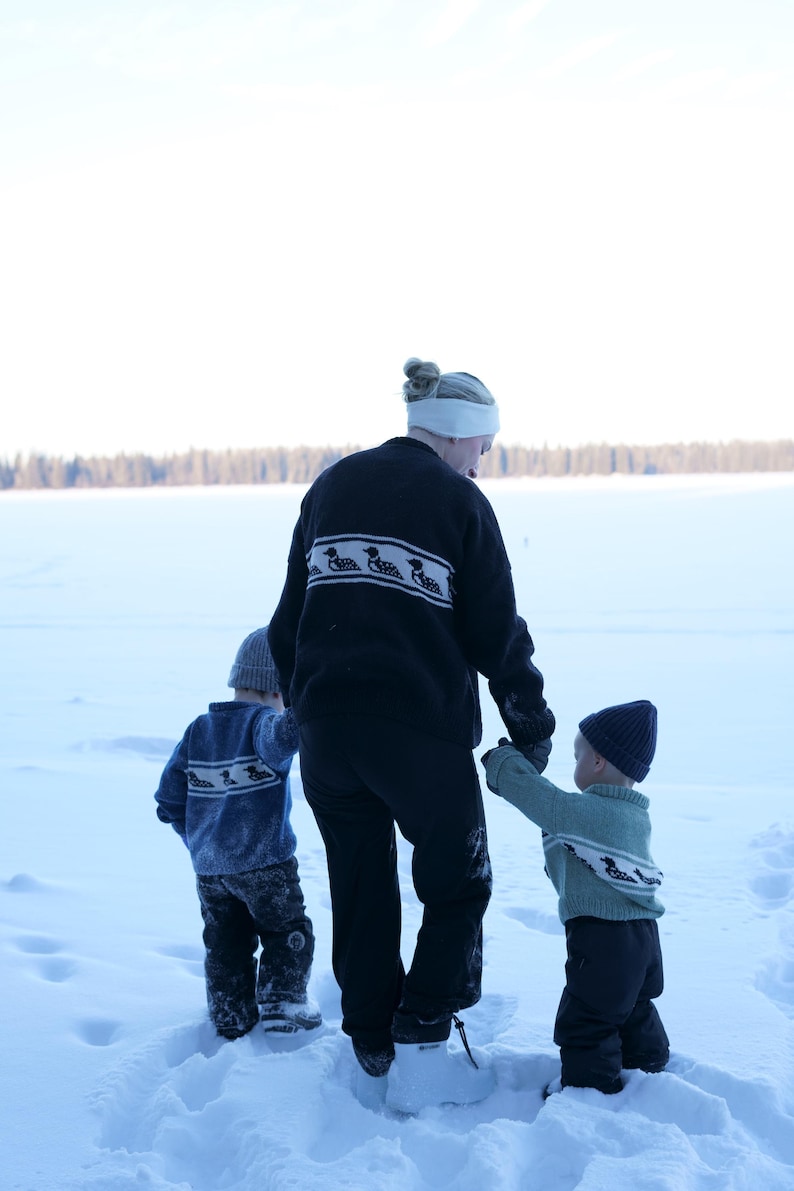 May include: A person and two children walking in the snow. The person wears a black sweater with a white and black pattern. The children wear sweaters and hats. The scene is set in a snowy landscape.