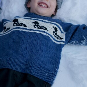 May include: A child wearing a blue sweater with a white band featuring black duck silhouettes, lying in the snow. The child is also wearing a gray hat and blue mittens. The sweater is a knit design.