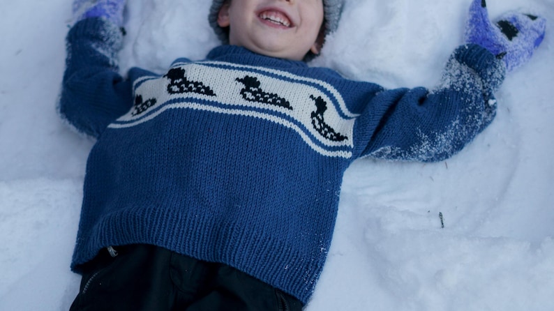 May include: A child wearing a blue sweater with a white band featuring black duck silhouettes, lying in the snow. The child is also wearing a gray hat and blue mittens. The sweater is a knit design.
