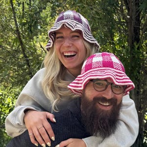 May include: Two crochet bucket hats featuring a checkered pattern. One hat is white and purple, while the other is white and bright pink. Both hats have a wide, slightly wavy brim and a rounded crown, crafted with a visible knit texture.