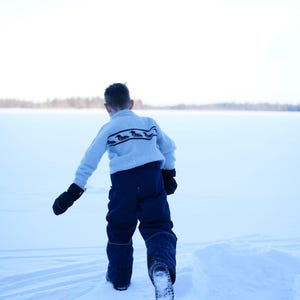 May include: A child in a light blue sweater with a dark pattern and navy blue snow pants walks across a snow-covered landscape. The child wears black mittens and boots, with a snowy background and a bright sky.