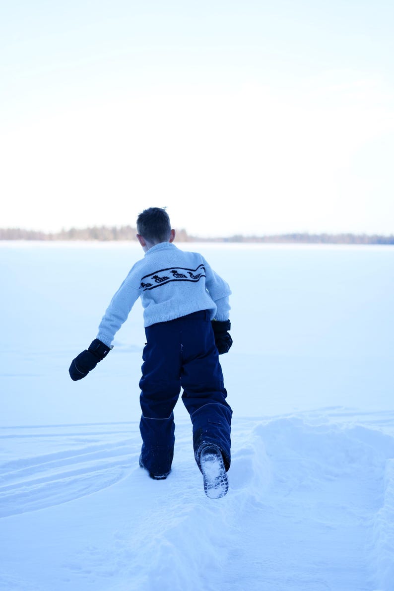 May include: A child in a light blue sweater with a dark pattern and navy blue snow pants walks across a snow-covered landscape. The child wears black mittens and boots, with a snowy background and a bright sky.