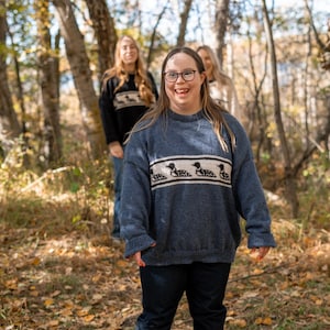 May include: A group of people wearing blue sweaters with a white band featuring a black duck design. The sweaters are paired with dark blue jeans. The background is a forest with trees and fallen leaves.