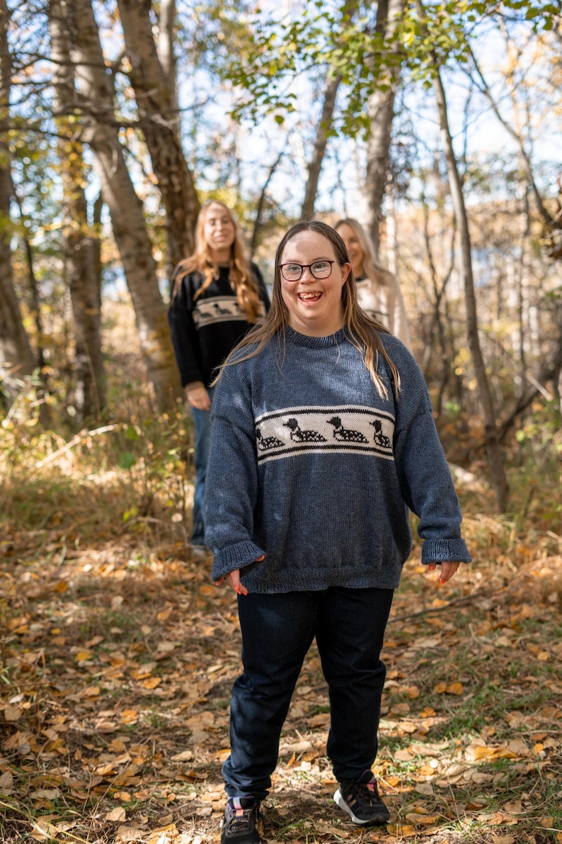 May include: A group of people wearing blue sweaters with a white band featuring a black duck design. The sweaters are paired with dark blue jeans. The background is a forest with trees and fallen leaves.