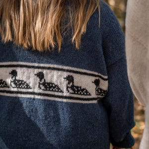 May include: A blue knit sweater with a white band featuring a black and white loon pattern. The sweater has long sleeves and a relaxed fit. The image shows the back of the sweater.