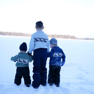 May include: Three children in winter clothing stand in the snow, facing away from the camera. They wear matching sweaters with a duck design, dark pants, and hats. The scene is set against a snowy landscape and a bright sky.
