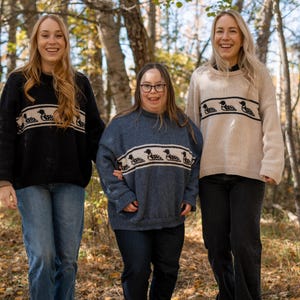 May include: Three people wearing sweaters with a horizontal band of loon designs. The sweaters are black, blue, and beige. They are wearing blue and black jeans. The background is a forest with trees and fallen leaves.