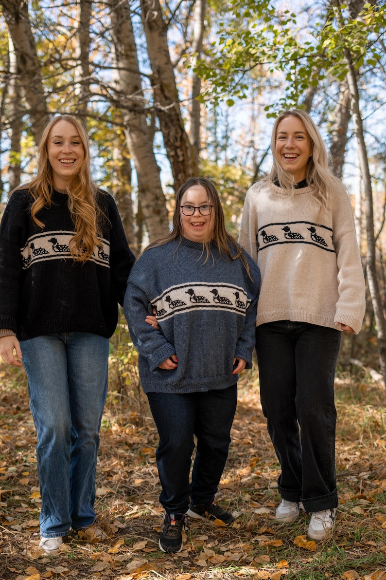 May include: Three people wearing sweaters with a horizontal band of loon designs. The sweaters are black, blue, and beige. They are wearing blue and black jeans. The background is a forest with trees and fallen leaves.
