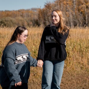 May include: Two people wearing matching sweaters outdoors. One sweater is blue, the other black. Both sweaters have a white band with a duck design. They are holding hands in a field of tall grass with trees in the background.