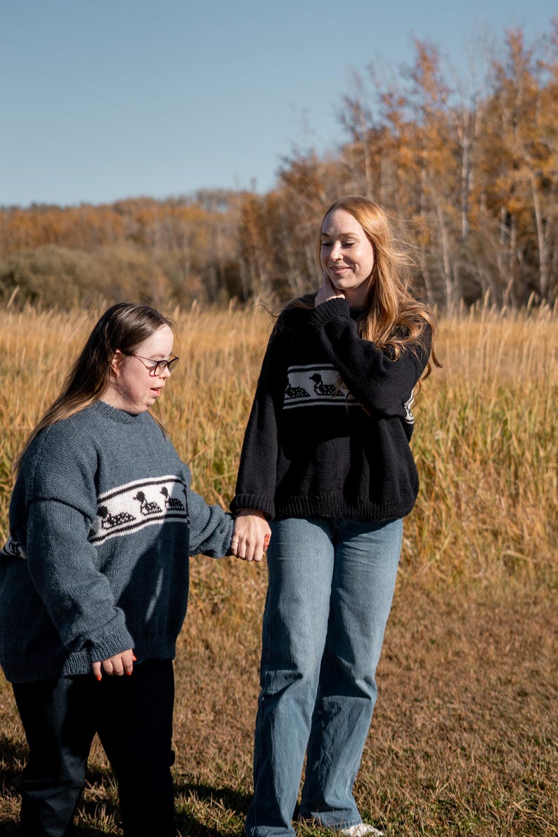 May include: Two people wearing matching sweaters outdoors. One sweater is blue, the other black. Both sweaters have a white band with a duck design. They are holding hands in a field of tall grass with trees in the background.