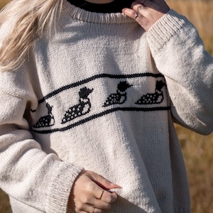May include: Cream-colored sweater with a black loon design. The sweater has a crew neck and long sleeves. The loon design is a horizontal band across the chest, featuring four loon silhouettes.
