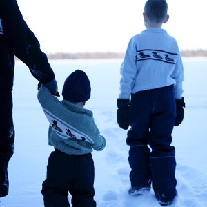 May include: Three children in winter clothing walking in the snow. The children are wearing blue and green sweaters with a duck design, dark blue pants, and black mittens. One child wears a blue knit cap.