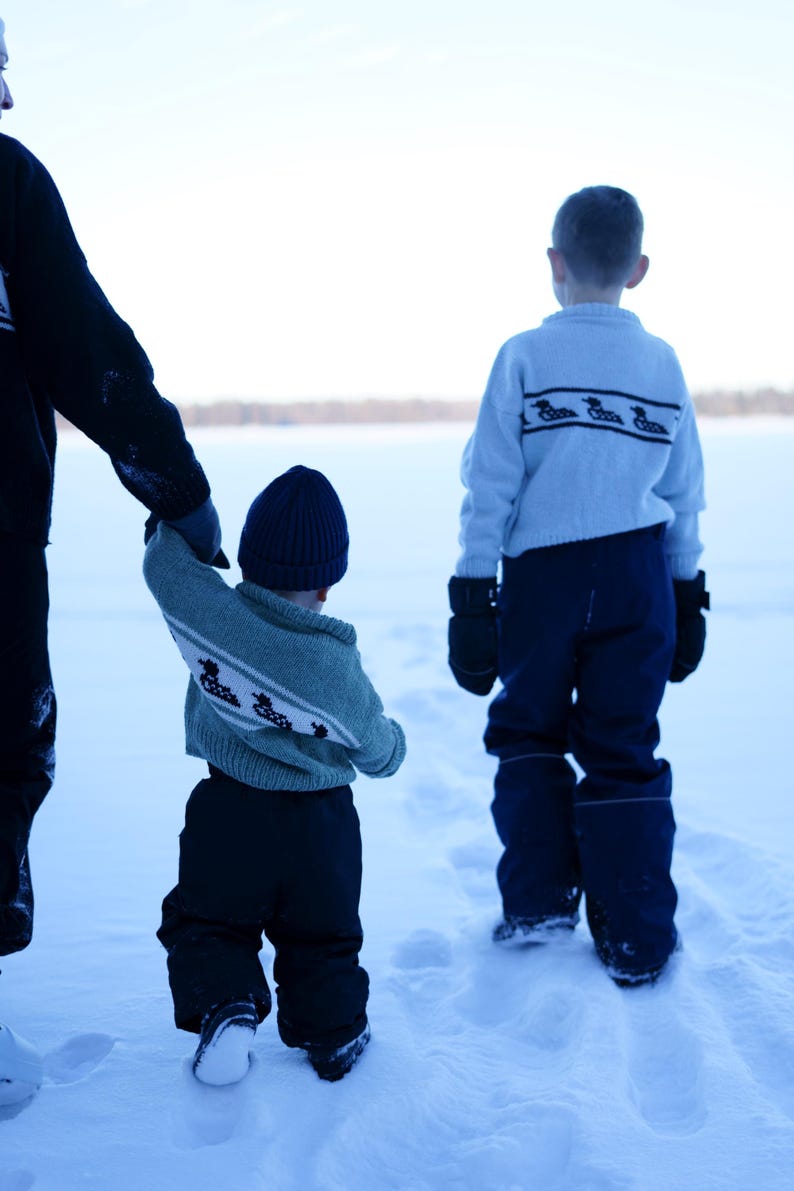 May include: Three children in winter clothing walking in the snow. The children are wearing blue and green sweaters with a duck design, dark blue pants, and black mittens. One child wears a blue knit cap.