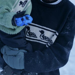 May include: A person wearing a navy blue sweater with a white band featuring a duck design. The person is holding a child wearing a green sweater with a similar duck pattern and blue mittens. The scene is outdoors in a snowy environment.
