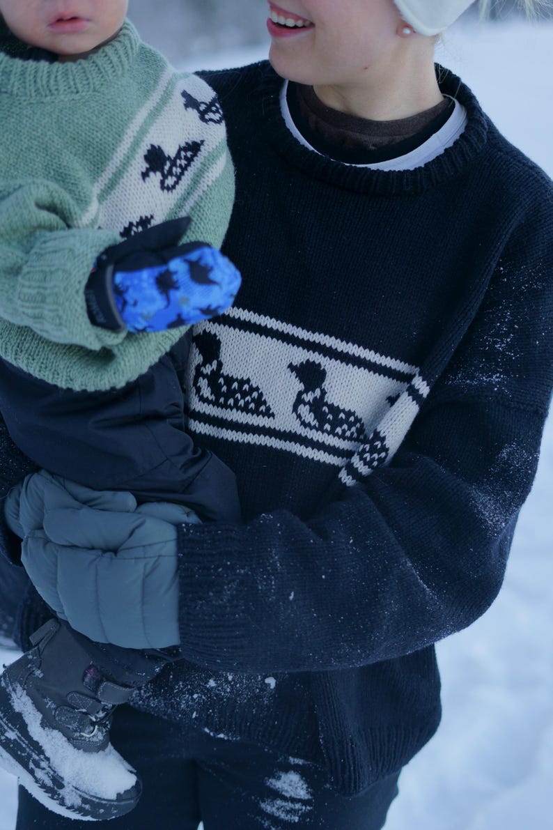 May include: A person wearing a navy blue sweater with a white band featuring a duck design. The person is holding a child wearing a green sweater with a similar duck pattern and blue mittens. The scene is outdoors in a snowy environment.