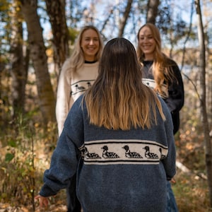 May include: A group of people wearing knitted sweaters outdoors. The front person wears a blue sweater with a horizontal band of black and white loon designs. The other two people wear cream and black sweaters with similar loon designs.