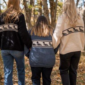 May include: Three people wearing sweaters with a loon design on the back, walking in a forest. The sweaters are black, blue, and cream-colored. The people are wearing jeans and sneakers. The background is a blur of trees and leaves.