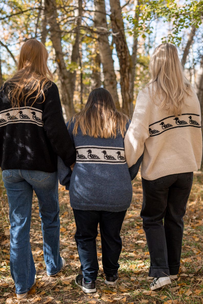 May include: Three people wearing sweaters with a loon design on the back, walking in a forest. The sweaters are black, blue, and cream-colored. The people are wearing jeans and sneakers. The background is a blur of trees and leaves.