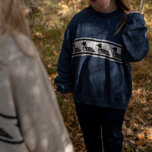 May include: A person wearing a dark blue knit sweater with a white and black loon design across the chest. The sweater is paired with black pants. The background shows a forest with fallen leaves.