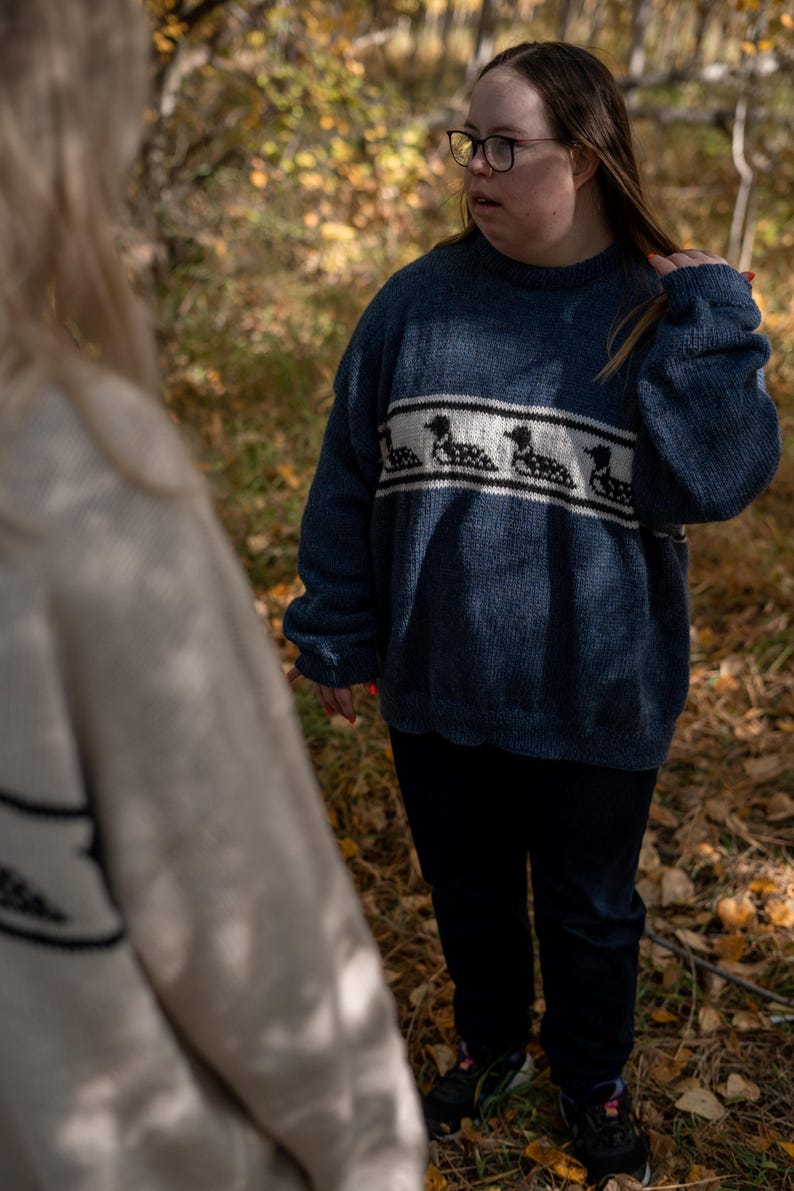 May include: A person wearing a dark blue knit sweater with a white and black loon design across the chest. The sweater is paired with black pants. The background shows a forest with fallen leaves.