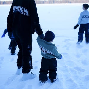 May include: A person and children walking in the snow. The person wears a black sweater with a white and black design, black pants, and white boots. A child wears a blue hat, green sweater with a white and blue design, and black pants.