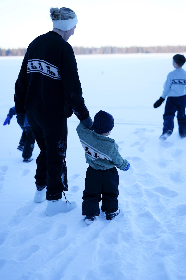 May include: A person and children walking in the snow. The person wears a black sweater with a white and black design, black pants, and white boots. A child wears a blue hat, green sweater with a white and blue design, and black pants.