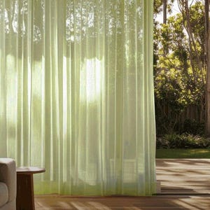 Puede incluir: Cortinas translúcidas de color verde claro cuelgan frente a una ventana grande, filtrando la luz solar. Las cortinas están plisadas verticalmente, creando un efecto suave y fluido. La habitación tiene un suelo de madera y una pequeña mesa auxiliar.