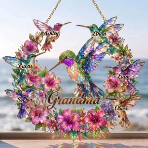 May include: A decorative hummingbird wreath with the word "Grandma" in the center. The wreath features colorful hummingbirds and flowers, with names like Ashley, Ethan, Lucas, Liam, Oliver, and Sophia. The wreath is suspended by a gold chain.