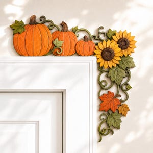 May include: A decorative corner door topper featuring three orange pumpkins of varying sizes, two yellow sunflowers, and green foliage with brown maple leaves. The design includes swirling vine accents and is shaped to fit the top corner of a door frame.