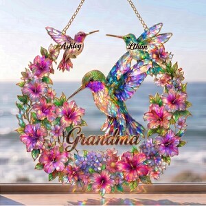 May include: A decorative hummingbird wreath with the names "Ashley" and "Ethan" on the birds. The word "Grandma" is written in cursive. The wreath is surrounded by pink, purple, and blue flowers, with a gold chain for hanging.